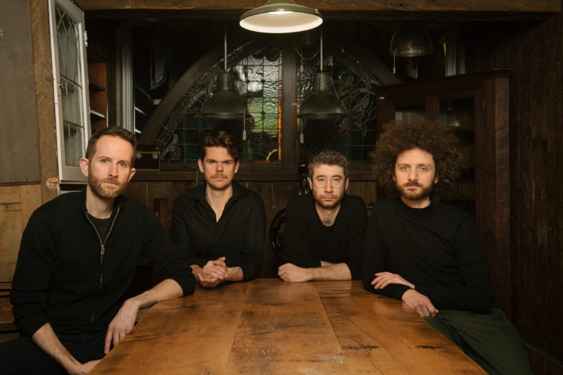 Four male members of a percussion ensemble are seated at a wooden table, with vibrant stained glass windows behind them. Each musician has a relaxed posture, as though reflecting their shared focus on music. The colorful stained glass adds a warm, artistic atmosphere to the scene.